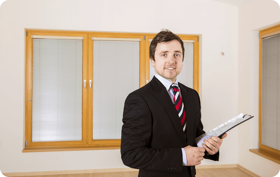 A suited man holds a clipboard in an empty room, appearing to assess the space around him.