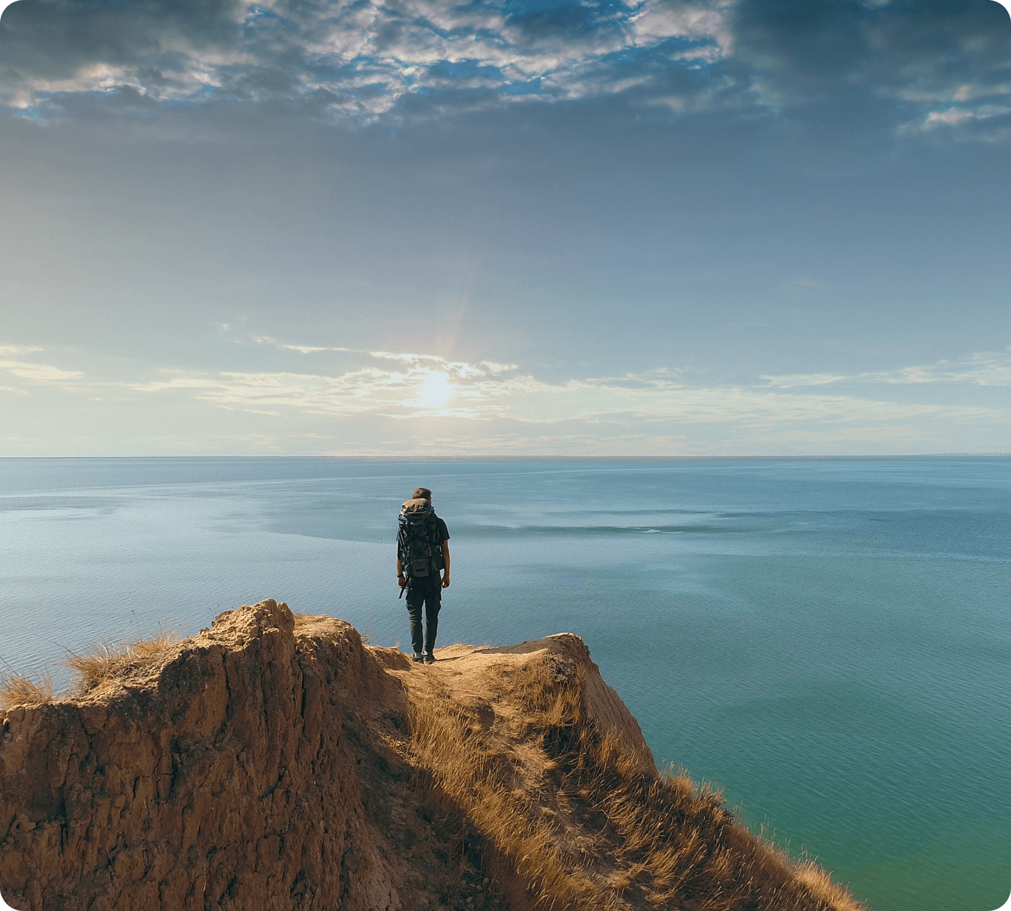 A person on a cliff edge looks over the ocean, surrounded by rugged terrain and a clear blue sky.